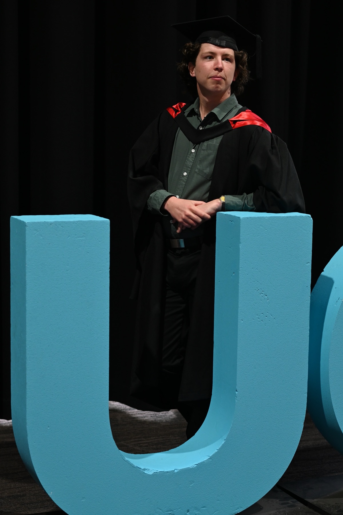 Josh in graduation robes, leaning on the giant blue letters of the University of Canberra sign
